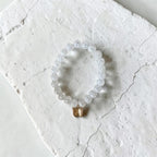 A beaded stretch bracelet made of clear, faceted round beads with a small gold-toned dangling charm, displayed on a textured white stone slab.