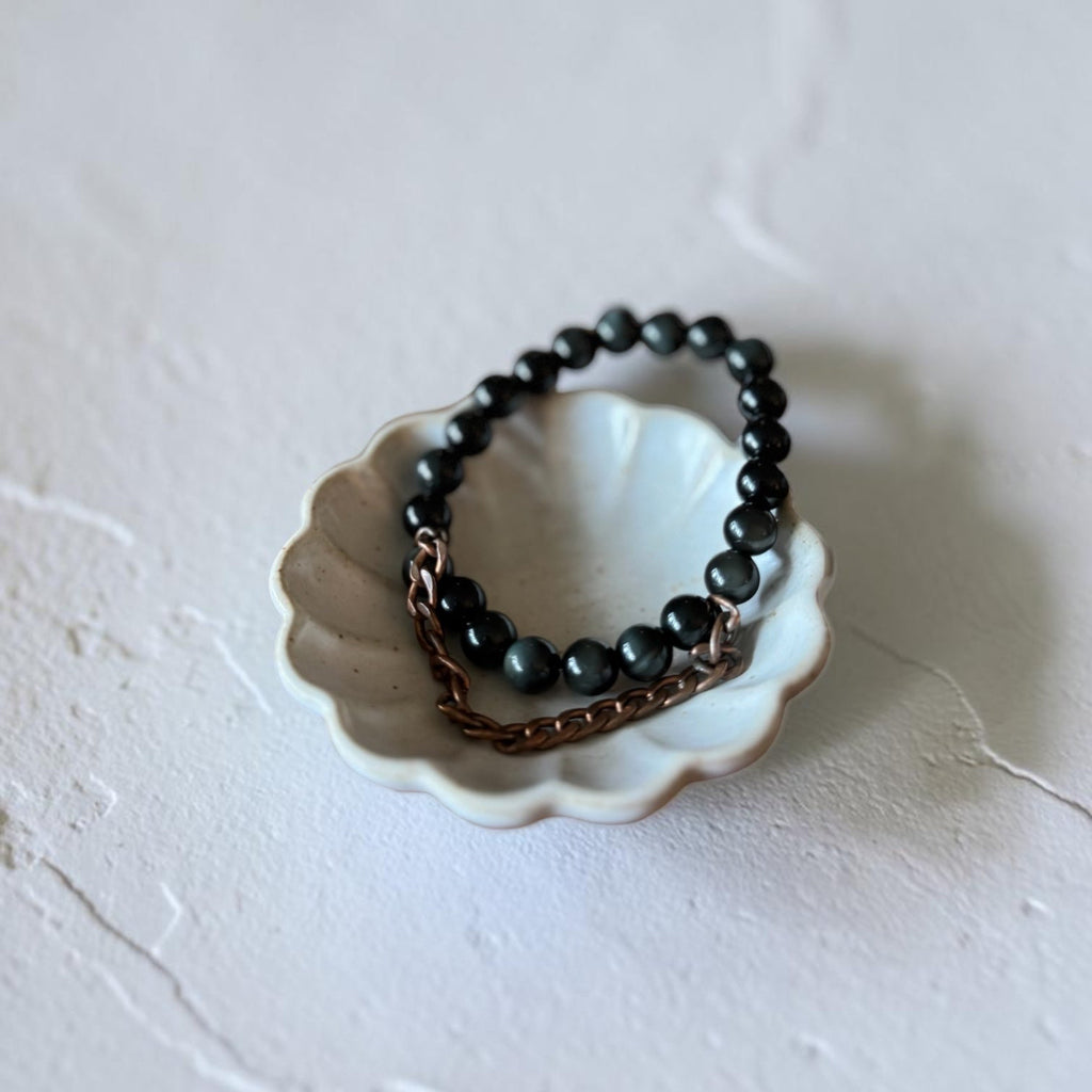 A beaded bracelet with dark gray beads and a copper chain link, displayed on a small white ceramic dish against a textured off-white background.