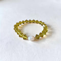 A beaded stretch bracelet featuring faceted olive-green beads and a single white pearl accent bead, displayed on a textured white surface.