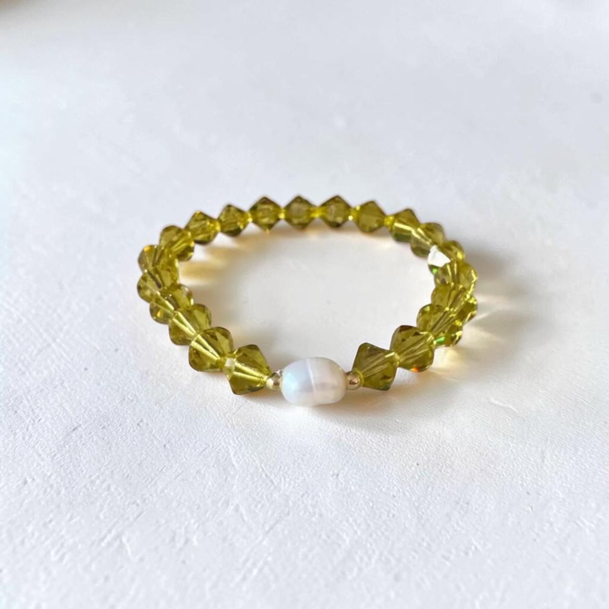 A beaded stretch bracelet featuring faceted olive-green beads and a single white pearl accent bead, displayed on a textured white surface.