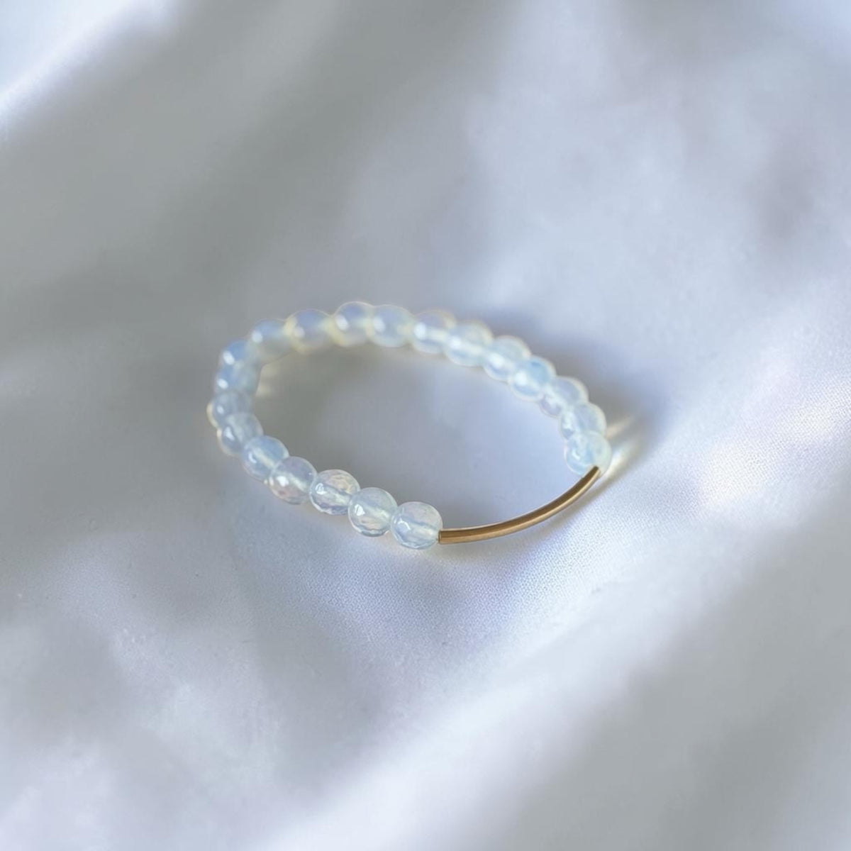 A beaded ring featuring translucent white and pale blue glass beads with a single gold metal segment, resting on a soft white fabric background.