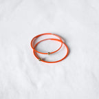 Two thin orange wire bracelets with small gold bead accents, arranged in a circle on a white textured fabric background.