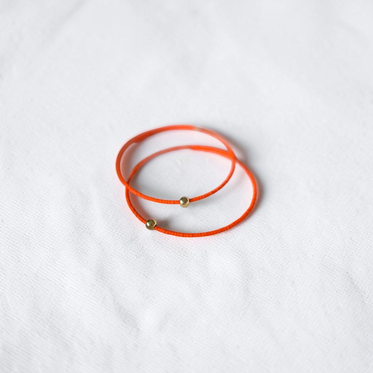 Two thin orange wire bracelets with small gold bead accents, arranged in a circle on a white textured fabric background.