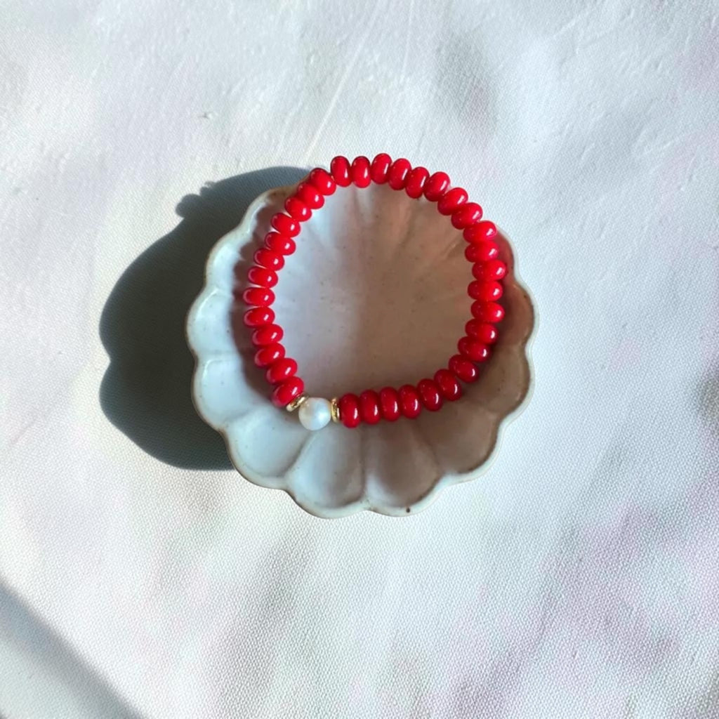 A red beaded bracelet with a single white pearl accent bead, displayed on a small white scalloped dish against a white fabric background.