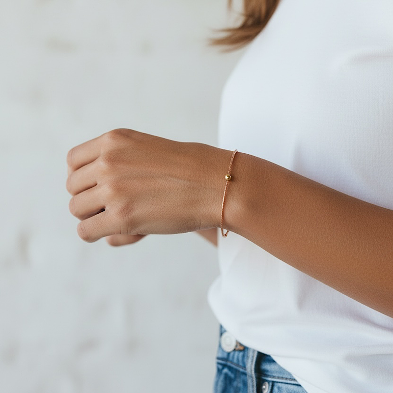 A delicate brown and gold bracelet with a small bead detail worn on a woman's wrist, shown in a lifestyle setting against a soft, light gray background.