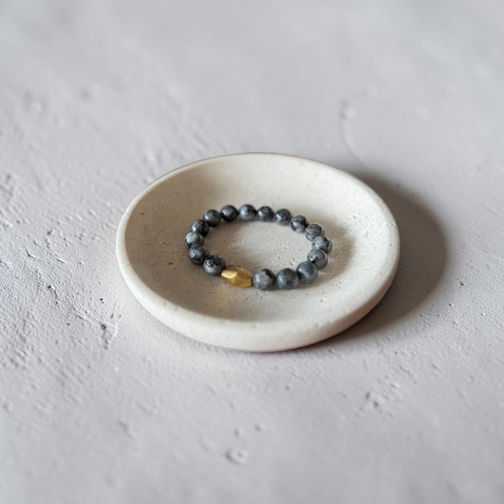 A beaded stretch bracelet with dark gray stone beads and a small gold accent bead, displayed on a small round ceramic dish against a textured light gray background.