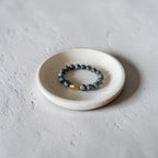 A beaded stretch bracelet with dark gray stone beads and a small gold accent bead, displayed on a small round ceramic dish against a textured light gray background.