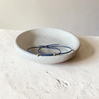 Two thin blue beaded bracelets with small gold beads, displayed inside a round textured off-white ceramic dish on a matching surface.