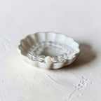 A beaded stretch bracelet with clear faceted beads and a single white pearl, displayed inside a small white ceramic dish on a textured light gray surface.