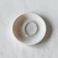 A small round ceramic dish with a speckled off-white glaze, holding a delicate beaded bracelet, set against a textured light gray concrete background.