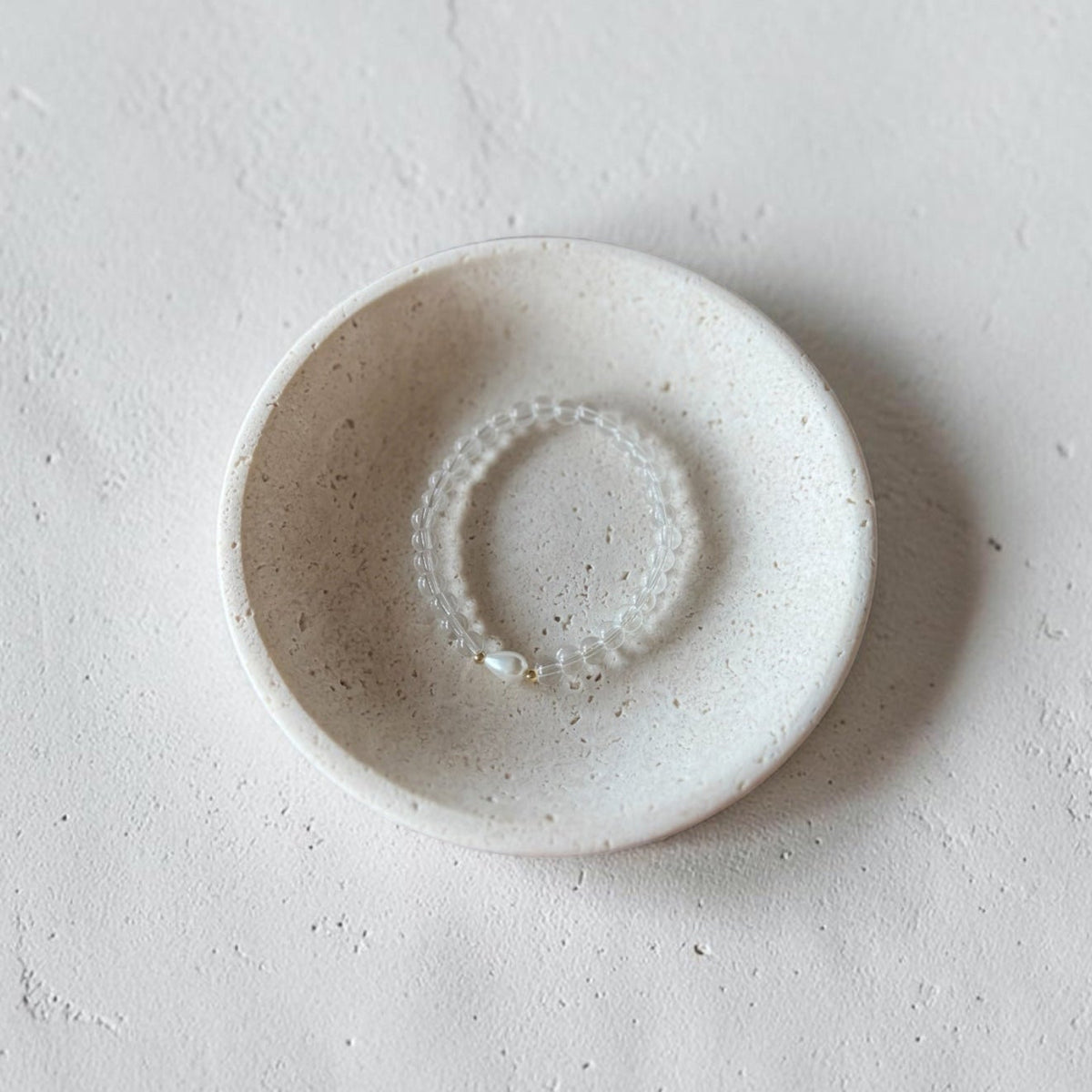 A small round ceramic dish with a speckled off-white glaze, holding a delicate beaded bracelet, set against a textured light gray concrete background.