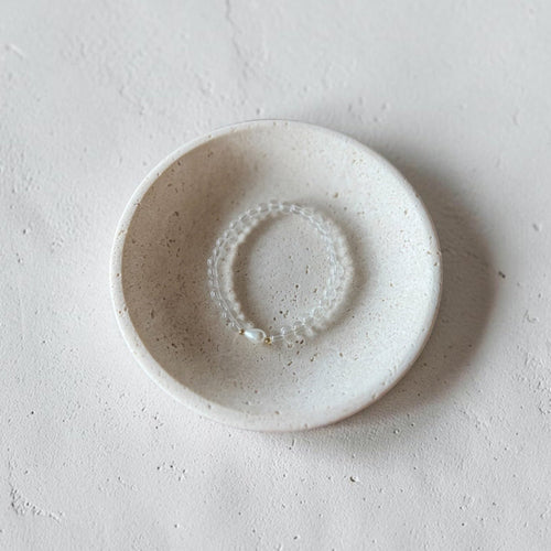 A small round ceramic dish with a speckled off-white glaze, holding a delicate beaded bracelet, set against a textured light gray concrete background.