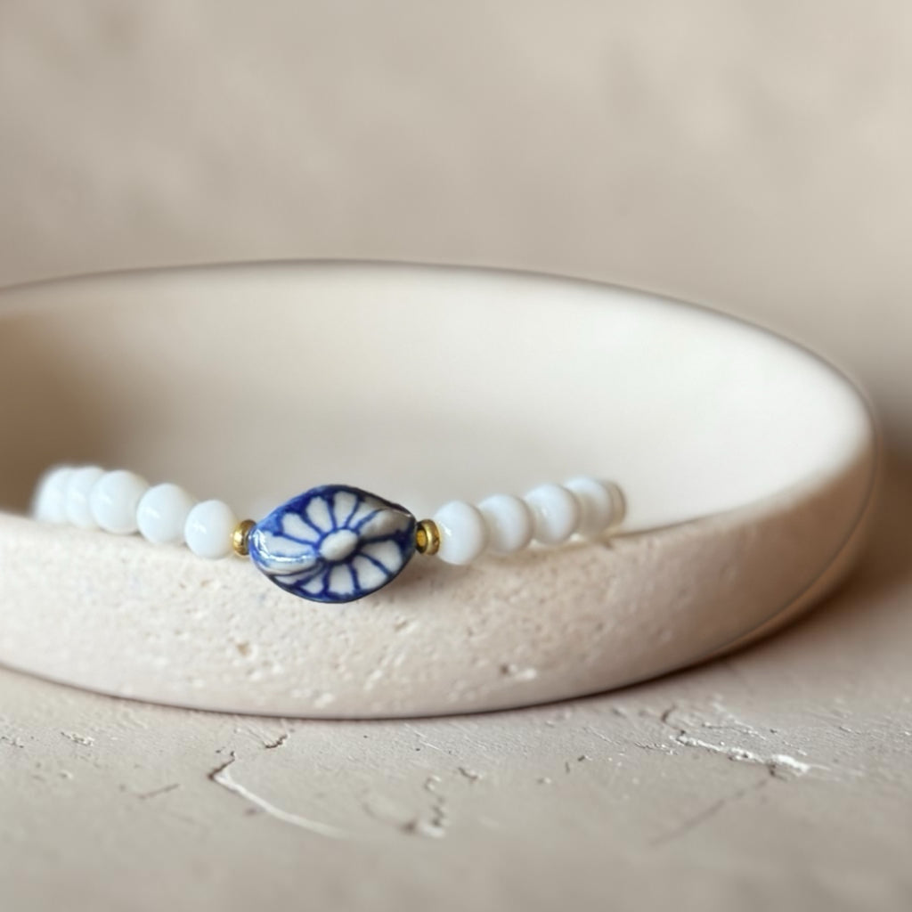 A beaded bracelet with white round beads and a central blue-and-white floral clover-shaped charm, displayed on a textured beige ceramic dish.