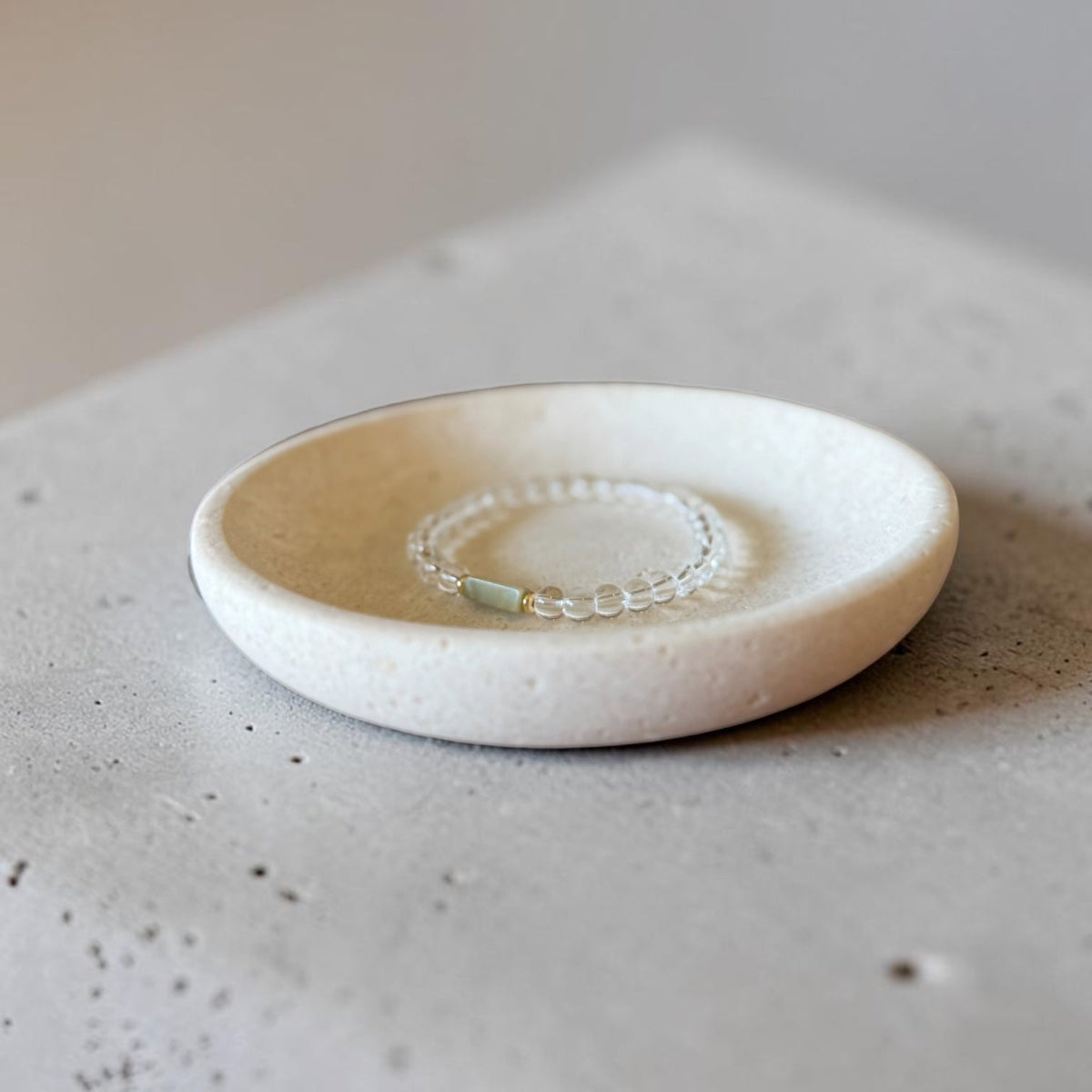 A delicate beaded bracelet with a light green stone accent resting inside a small, round, textured off-white ceramic dish, set on a concrete surface.