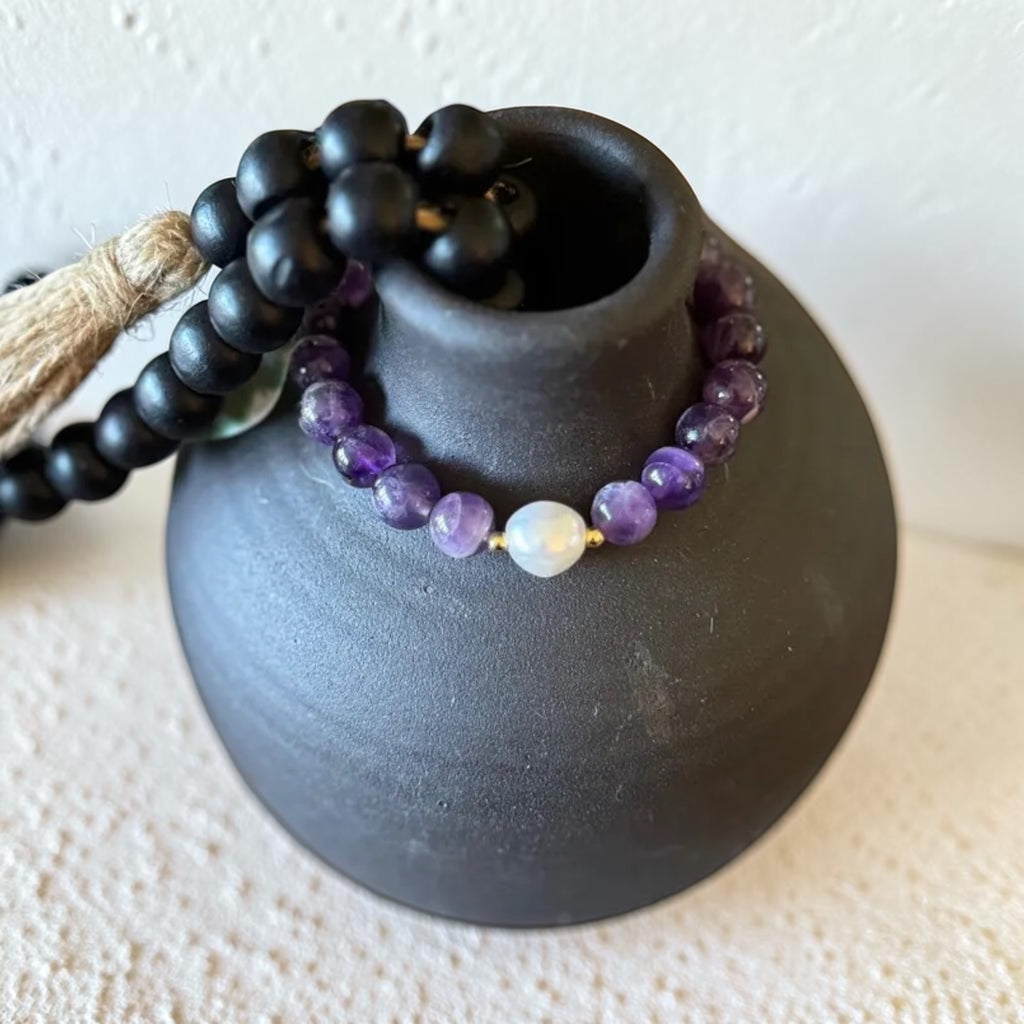 A beaded bracelet with purple amethyst and white moonstone beads draped over a matte black ceramic vase, with a black beaded strand and tan cord visible behind it on a textured cream surface.