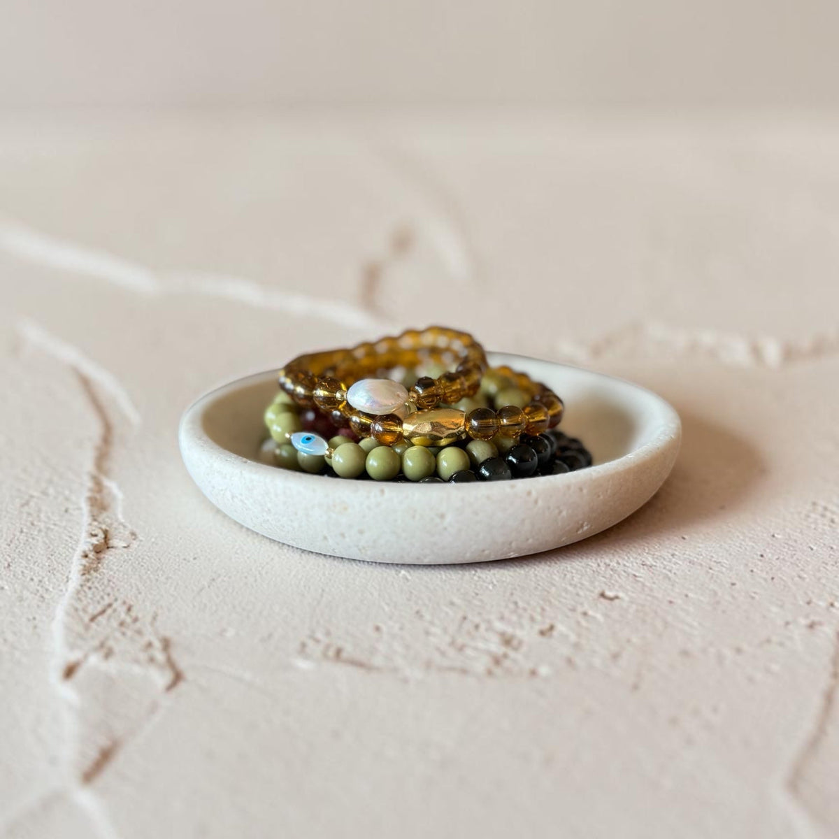 A small round white ceramic jewelry tray holding several beaded bracelets, including green, black, and amber tones, set against a textured beige background.