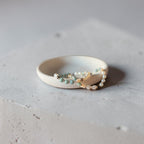 A delicate beaded bracelet with white pearls, light blue beads, and small gold-toned beads rests inside a matte off-white ceramic ring holder on a textured light gray concrete surface.