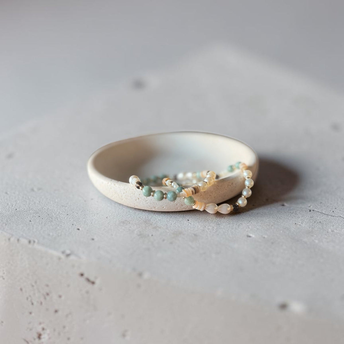 A delicate beaded bracelet with white pearls, light blue beads, and small gold-toned beads rests inside a matte off-white ceramic ring holder on a textured light gray concrete surface.