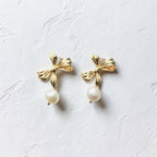 A pair of gold bow stud earrings with dangling white pearls, displayed on a textured white surface.