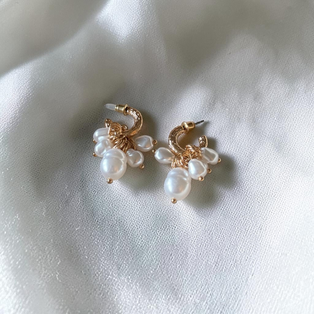 A pair of gold hoop earrings with clusters of white freshwater pearls, displayed on a soft, draped white fabric background.