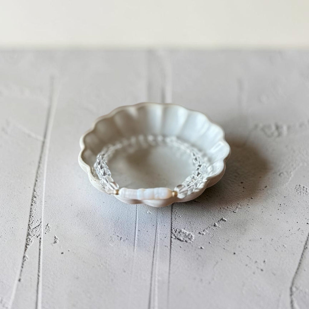 A small scalloped ceramic dish with a white floral pattern and a beaded inner rim, holding a delicate clear beaded bracelet, set on a textured light gray surface.