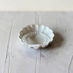 A small scalloped ceramic dish with a white floral pattern and a beaded inner rim, holding a delicate clear beaded bracelet, set on a textured light gray surface.