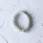 A white beaded stretch bracelet with a textured, scalloped edge and a single larger pearl-like bead at the front, displayed on a textured white surface.
