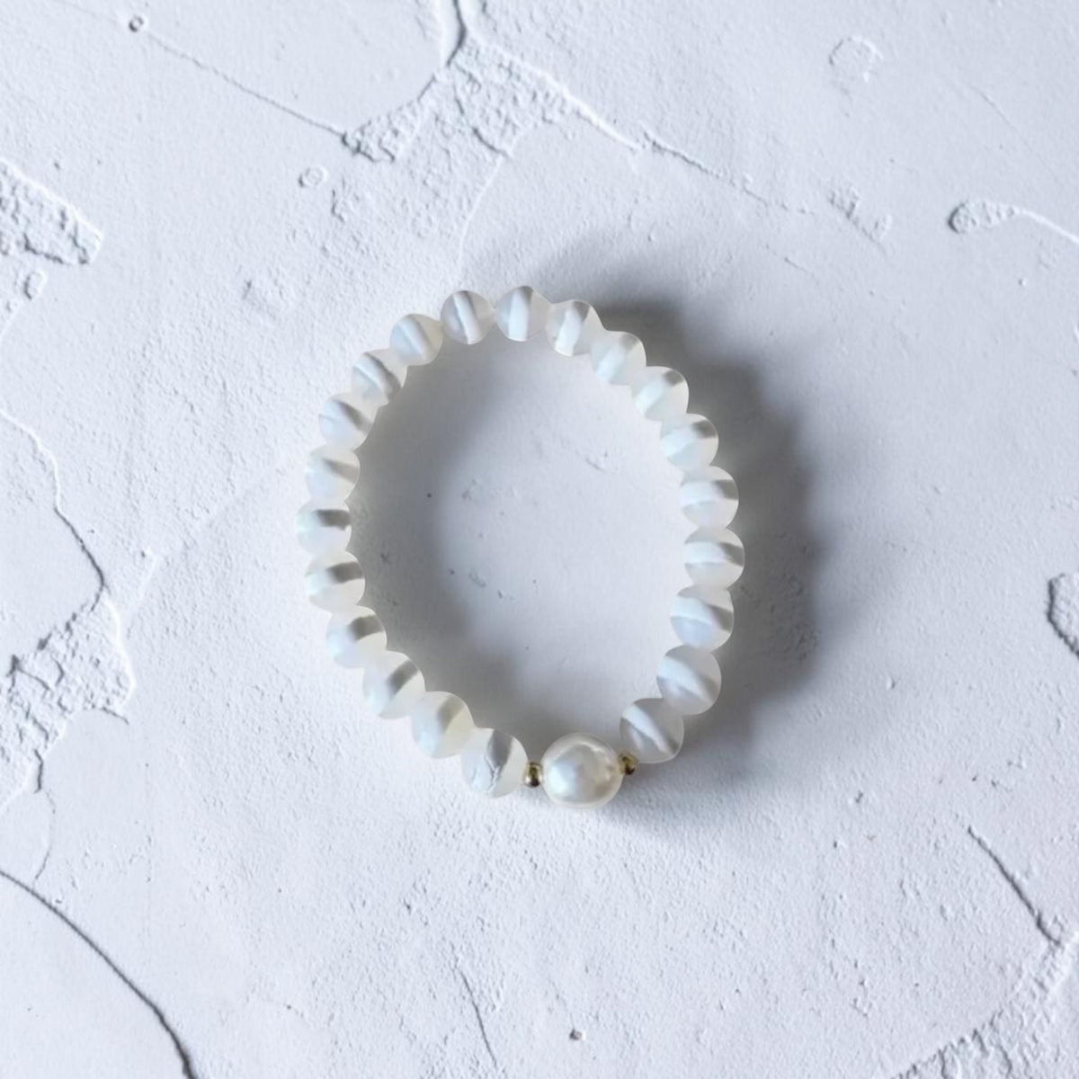 A white beaded stretch bracelet with a textured, scalloped edge and a single larger pearl-like bead at the front, displayed on a textured white surface.