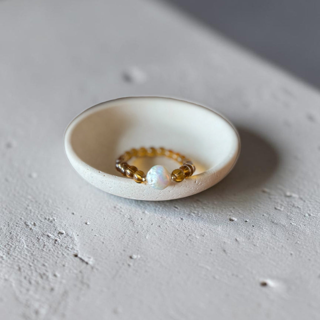 A small beaded bracelet with amber-brown beads and a white pearl-like center, displayed inside a shallow white ceramic dish on a textured light gray surface.
