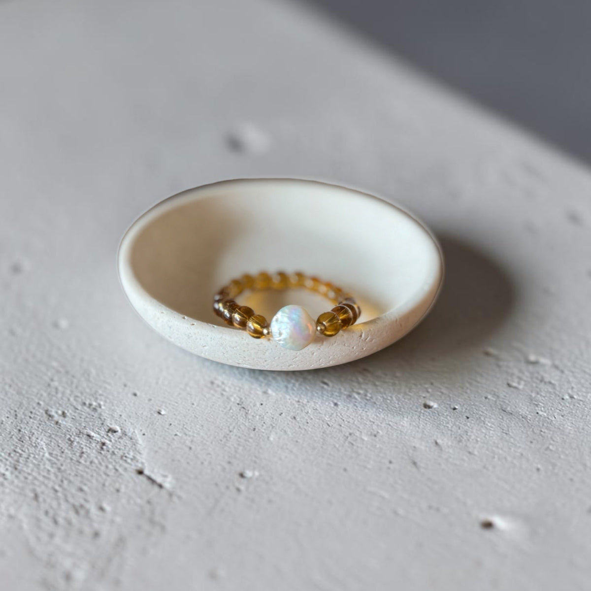 A small beaded bracelet with amber-brown beads and a white pearl-like center, displayed inside a shallow white ceramic dish on a textured light gray surface.