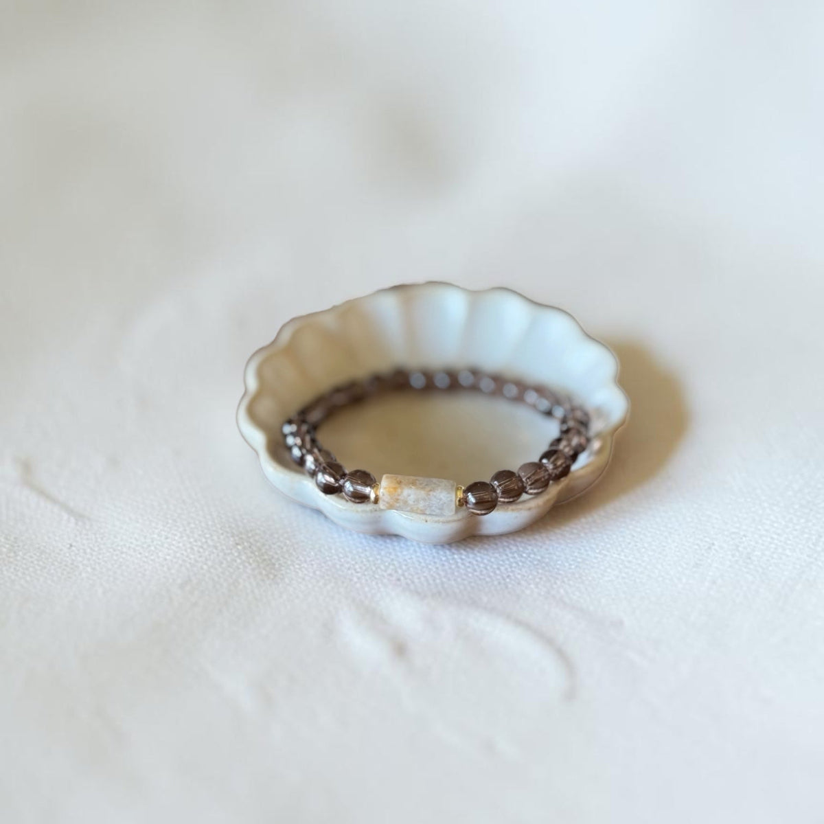 A beaded stretch bracelet with dark brown beads and a central cream-colored stone, displayed inside a small white ceramic dish on a soft, textured white fabric background.