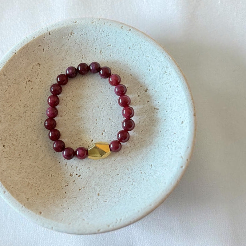 A beaded stretch bracelet with deep burgundy red round beads and a faceted gold accent bead, displayed inside a light beige ceramic dish on a white fabric background.