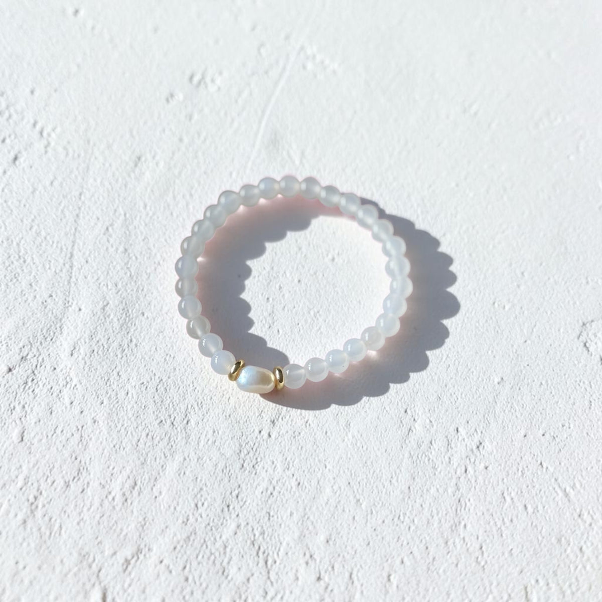 A beaded stretch ring featuring translucent white beads and a small gold-toned accent bead, photographed on a textured white surface with a soft shadow.