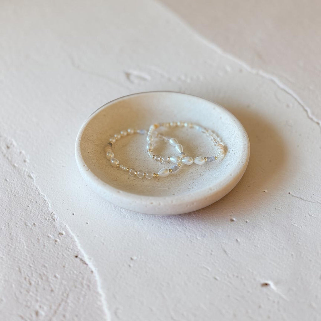 A pair of delicate pearl beaded bracelet resting on a small, round, shallow ceramic dish, set against a textured, light beige plaster background.