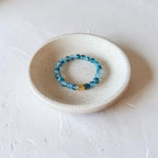 A beaded stretch bracelet with light blue translucent beads and a small gold accent, displayed inside a small speckled ceramic dish on a textured white surface.