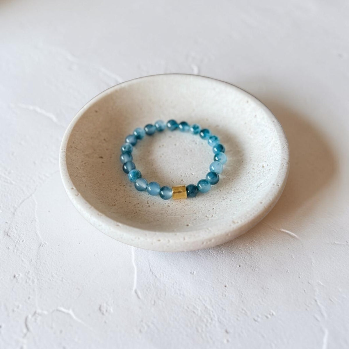 A beaded stretch bracelet with light blue translucent beads and a small gold accent, displayed inside a small speckled ceramic dish on a textured white surface.