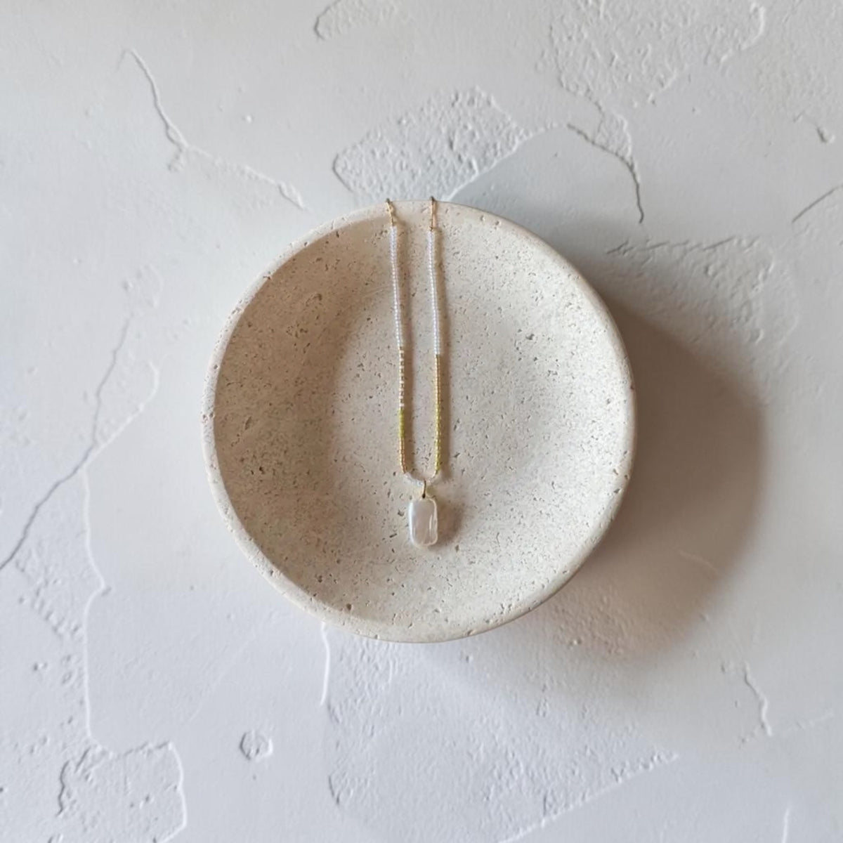 A handmade ceramic jewelry dish in a speckled cream-beige glaze, featuring a vertical slit down the center holding a small white bead, photographed from a top-down angle on a textured white surface.
