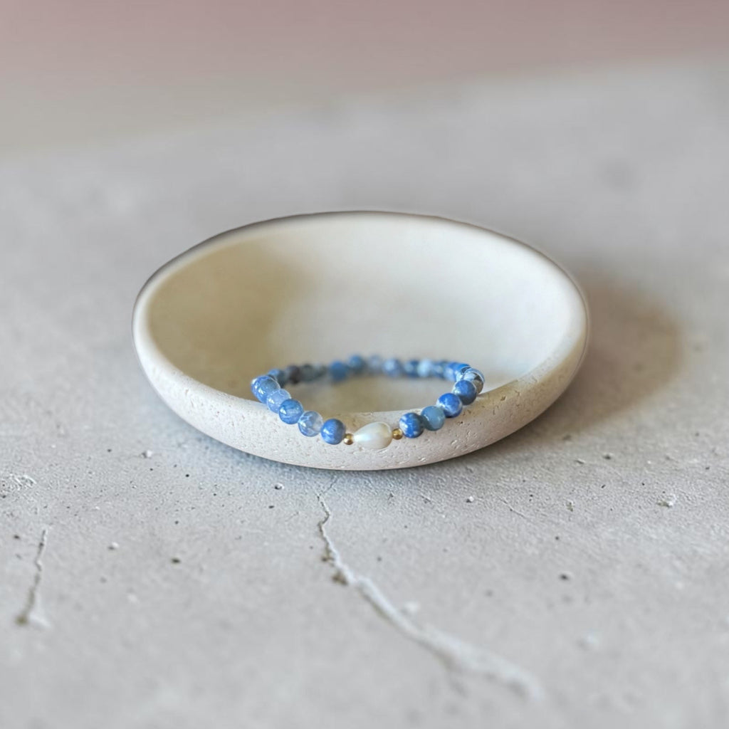 A beaded bracelet with light blue and white beads rests inside a small, shallow, off-white ceramic dish on a textured gray surface.