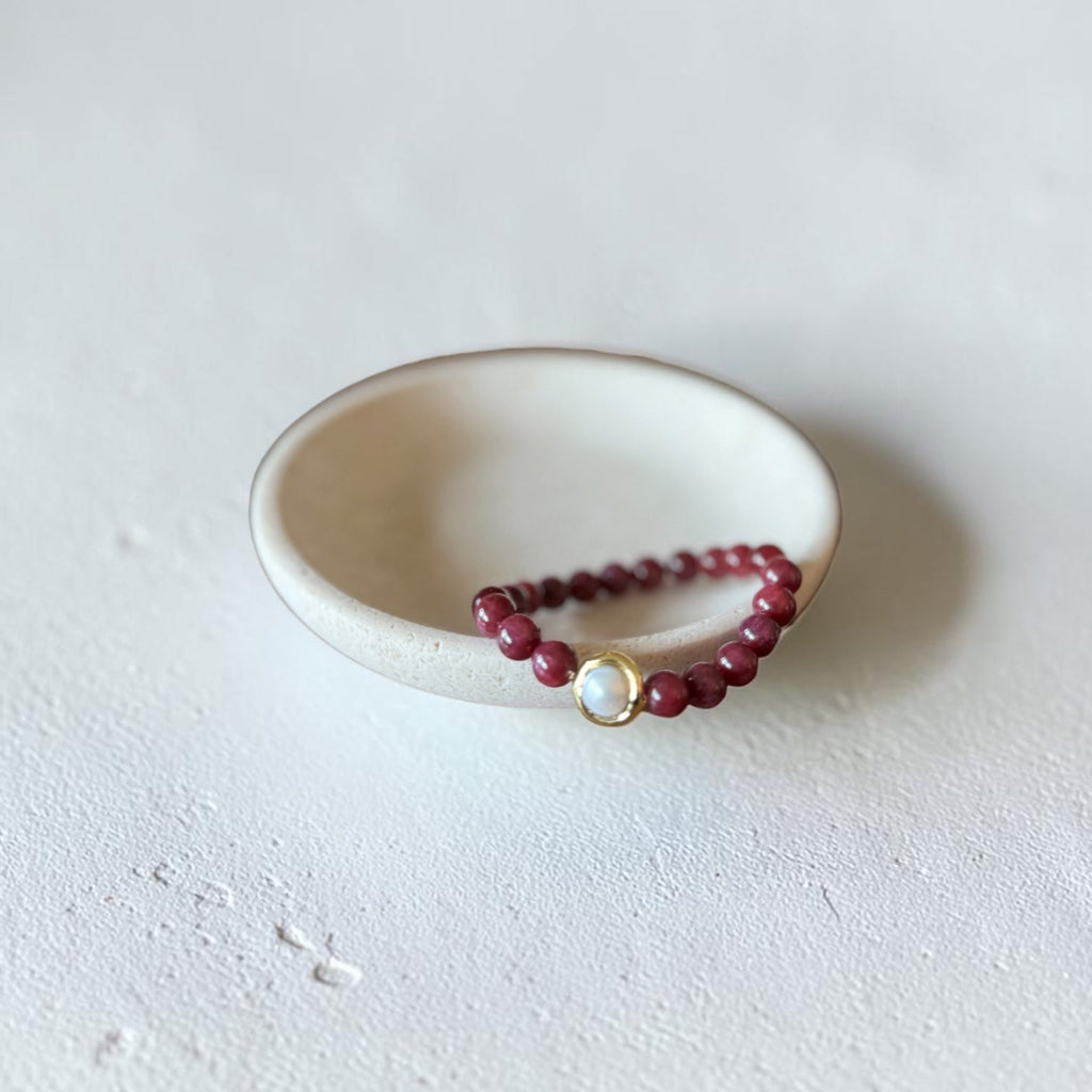 8mm Red Garnet Jade Bead Bracelet with a Gold And Pearl Accent. Accent has a round pearl with gold surrounding the pearl itself. This bracelet is sitting in a dish.