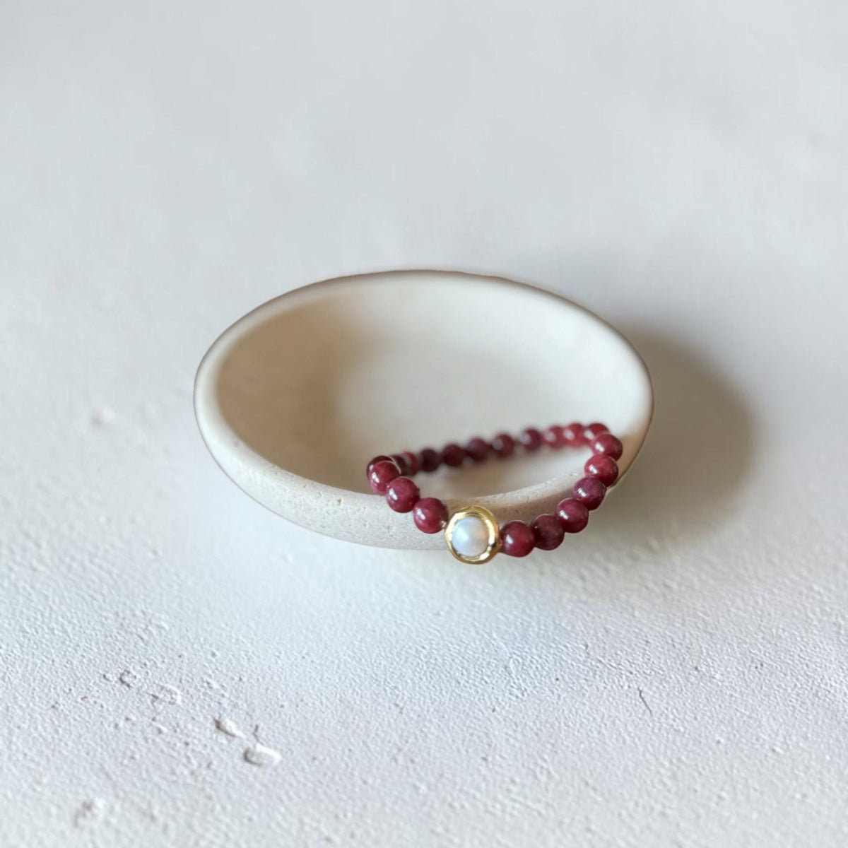 8mm Red Garnet Jade Bead Bracelet with a Gold And Pearl Accent. Accent has a round pearl with gold surrounding the pearl itself. This bracelet is sitting in a dish.