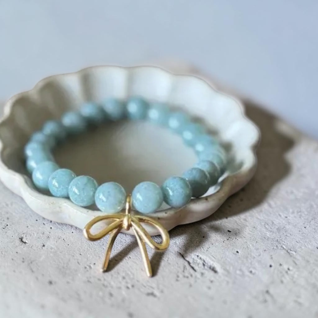A light blue beaded bracelet with a gold bow charm, displayed on a small white ceramic dish over a textured gray surface.