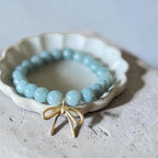 A light blue beaded bracelet with a gold bow charm, displayed on a small white ceramic dish over a textured gray surface.