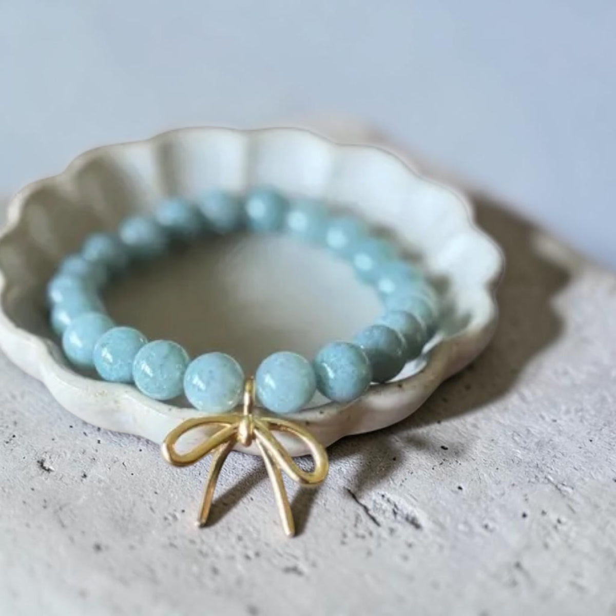 A light blue beaded bracelet with a gold bow charm, displayed on a small white ceramic dish over a textured gray surface.