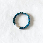 A beaded stretch bracelet with dark teal, blue, and green beads, featuring a small white stone accent, displayed on a textured white background.