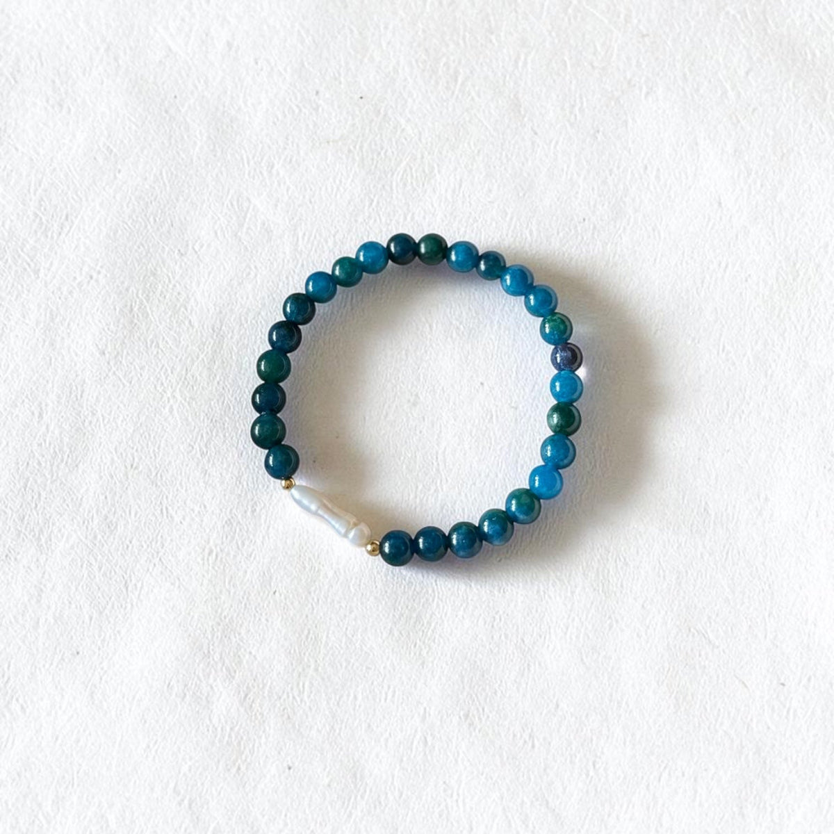 A beaded stretch bracelet with dark teal, blue, and green beads, featuring a small white stone accent, displayed on a textured white background.