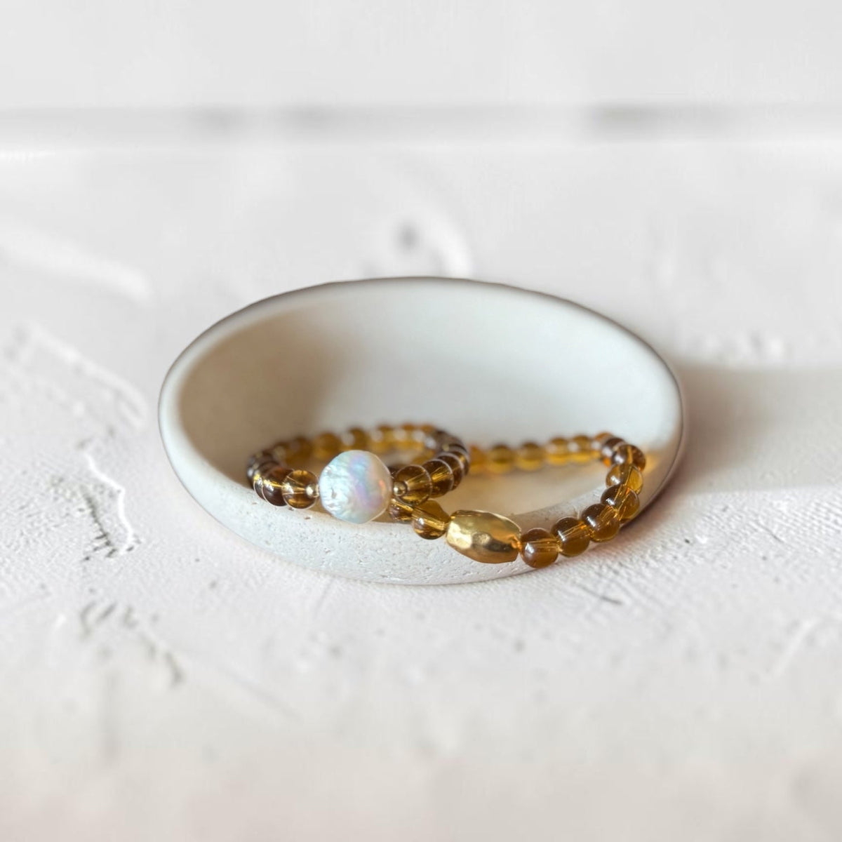 Two beaded stretch bracelets displayed in a small white ceramic dish. One bracelet features translucent amber-brown beads, while the other is made of similar amber beads with a single round white opal-like bead.