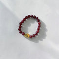 A beaded stretch bracelet made of dark red round beads with a faceted gold accent bead, displayed on a textured white surface with soft shadows.