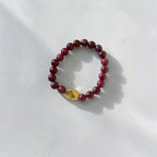 A beaded stretch bracelet made of dark red round beads with a faceted gold accent bead, displayed on a textured white surface with soft shadows.