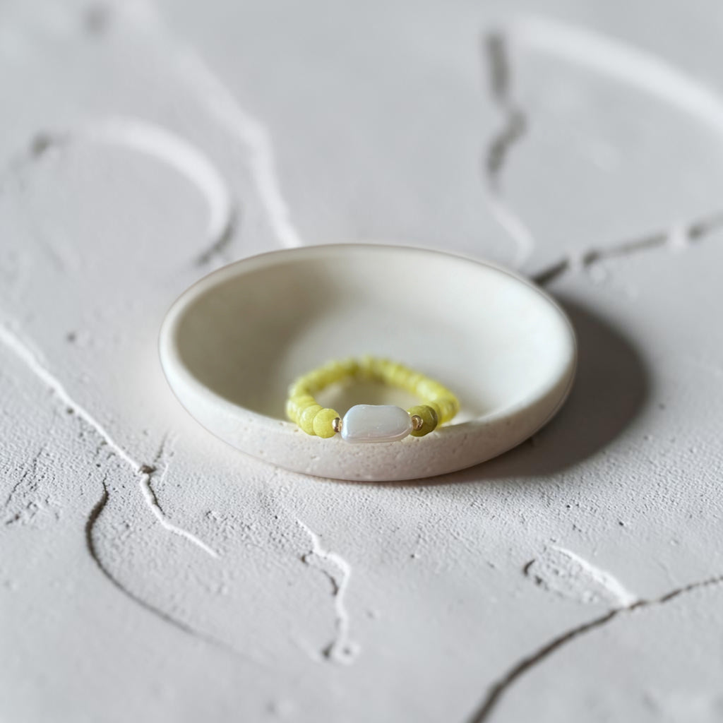 A yellow beaded bracelet with a white oval stone, displayed inside a small white ceramic dish on a textured light gray surface.
