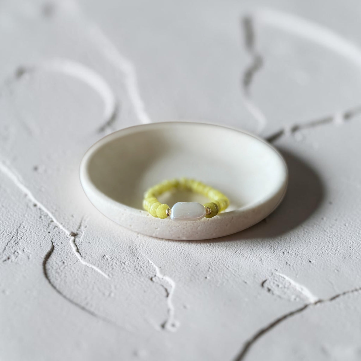 A yellow beaded bracelet with a white oval stone, displayed inside a small white ceramic dish on a textured light gray surface.